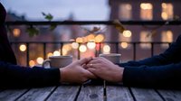 Romantic couple holding hands across wooden table on evening terrace with warm bokeh lights creating intimate atmosphere for date night celebration.