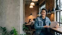 Young Asian man working on laptop in modern industrial cafe with brick walls and pendant lighting for remote work and freelancing concepts.