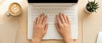 Overhead view of female hands typing on a white laptop at a wooden desk with coffee, notebook, and succulent plant.