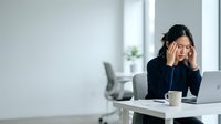 Asian female professional in dark business attire experiencing work stress at office desk with laptop and coffee cup, suitable for corporate wellness and burnout content.