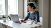 Middle-aged Hispanic woman working on laptop at home office desk with coffee cup and plants, remote work lifestyle for business productivity.