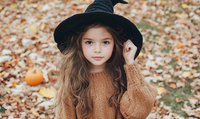 Young girl in black witch hat and cozy knit sweater poses among autumn leaves and pumpkins, capturing magical Halloween spirit with natural fall backdrop.