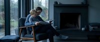 Young Caucasian woman sitting in armchair reading book by fireplace in modern living room on rainy day, cozy indoor atmosphere.