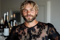 Confident Caucasian man with beard and blonde wavy hair wearing black lace transparent shirt, smiling at camera against blurred bar background with wine bottles.