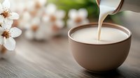 Pouring cream or milk into ceramic bowl with spring cherry blossoms in background on wooden surface, creating soft and serene morning atmosphere.
