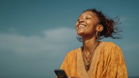 Young African American woman laughing joyfully outdoors with phone in hand against blue sky background, expressing happiness and positive emotions.