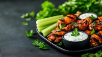 Grilled buffalo chicken wings served with celery sticks and ranch dipping sauce on black plate. Fresh herbs garnish adds vibrant color to spicy appetizer.