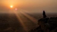 Silhouette of person standing on rocky cliff overlooking vast desert landscape during golden sunset with dramatic sun rays piercing through misty atmosphere.