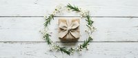 Kraft paper gift box with natural ribbon surrounded by delicate white flowers and green foliage wreath on rustic white wooden background for celebrations.