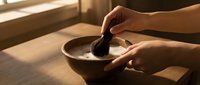 Hands preparing traditional matcha tea with bamboo whisk in wooden bowl on table near window with warm natural sunlight streaming across surface.