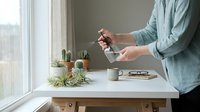 Woman in mint green shirt using smartphone at white desk with air plants and coffee cup near window for modern lifestyle and technology concept.