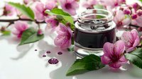 A glass jar filled with dark liquid surrounded by fresh pink flowers and green leaves on a white surface, suitable for cosmetics, skincare, wellness, and beauty product marketing.