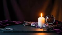 Glowing white candle with amethyst crystals, lavender sprigs, and a crystal ball on dark slate, surrounded by purple fabric.