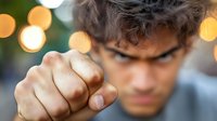 Young Hispanic male pointing finger at camera with intense expression and bokeh lights background, creating dramatic confrontational perspective.
