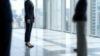 Businesswoman in formal black suit standing in modern high-rise office with floor-to-ceiling windows overlooking city skyline, professional corporate environment.