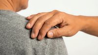 Hand gently placed on shoulder of elderly person in gray sweater, symbolizing support, empathy, and care on light background.