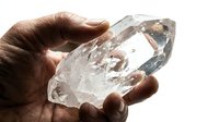 Hand of a dark-skinned man holding a large clear quartz crystal point, showcasing natural mineral beauty against a light background.