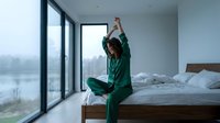 Woman in green pajamas stretching on bed by large window overlooking misty winter landscape. Morning wellness and relaxation routine for lifestyle and home decor content.