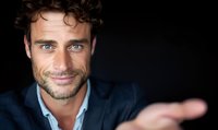 Confident Caucasian businessman with bright blue eyes and charming smile gestures while wearing navy suit jacket against dark background, professional headshot style portrait.