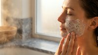 Young woman applying facial cleanser with foam bubbles during morning skincare routine in bright bathroom with natural window light for wellness and beauty.
