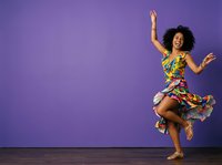 Young Black woman with curly hair wearing colorful floral dress, dancing joyfully with arms raised against purple background, barefoot on wooden floor for lifestyle and celebration content.