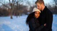 Interracial couple embracing in snowy winter park during golden hour sunset. Romantic moment between African American woman and Caucasian man outdoors.