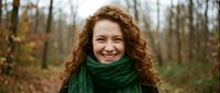 Happy young Caucasian woman with curly red hair wearing green knit scarf smiling outdoors in autumn forest for lifestyle and seasonal content.