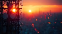 Silhouette of telecommunications tower against dramatic orange sunset sky with bokeh lights and urban cityscape in background creating atmospheric scene.