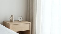 Wooden nightstand with drawer next to bed, featuring ceramic mug and analog clock on top, minimal Scandinavian bedroom interior design.