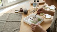 Crafting resin art with pressed flowers and fern leaves, a person arranges botanicals in a mold on a wooden table near a bright window.