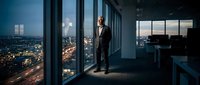 Businessman in formal suit standing in modern high-rise office at night, overlooking illuminated cityscape through floor-to-ceiling windows, professional corporate environment.