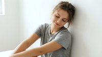 Young Caucasian woman in gray t-shirt sitting peacefully with eyes closed, expressing contentment and relaxation in bright indoor setting for wellness concepts.