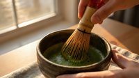 Matcha tea preparation with traditional bamboo whisk in ceramic bowl, whisking green powder into hot water for ceremonial beverage making and wellness rituals.