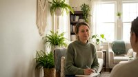 Middle-aged Caucasian woman in green sweater sitting in modern living room with houseplants, natural lighting, cozy home interior design concept.
