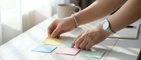 Woman's hands arranging colorful sticky notes on white desk near coffee cup, organizing tasks and planning workflow for productivity and time management.