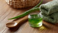Fresh aloe vera gel in glass jar with wooden spoon and green towels on rustic wooden table, natural skincare and wellness concept.