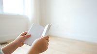 Person with medium skin tone holding open blank white book or notebook indoors near window with natural light, suitable for education, reading, publishing, and business concepts.