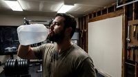 Middle-aged man with dark beard and hair, wearing casual tan shirt, drinking water from clear bottle in workshop garage setting with tools and equipment visible in background.