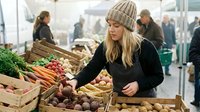 Young blonde woman in knit beanie shopping for fresh organic vegetables at bustling farmers market with wooden crates full of seasonal produce.