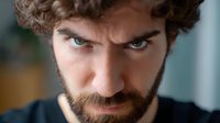Close up portrait of young bearded Caucasian man with intense gaze and serious expression looking directly at camera against blurred background. Dramatic lighting emphasizes facial features.