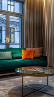 Modern living room interior with emerald green velvet sofa, orange accent pillows, marble coffee table and floor-to-ceiling windows. Luxurious urban apartment design.