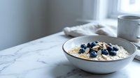 Bowl of oatmeal topped with blueberries and almonds on marble table with coffee cup and pillows in bright morning light.