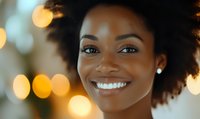 Young African American woman with natural afro hair smiling warmly against blurred golden bokeh lights background, expressing joy and confidence in casual setting.
