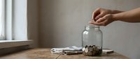 Hand placing coins into glass jar on wooden table near window for savings and financial planning goals.