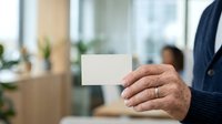 Mature man in business casual attire holding blank white card in modern office environment with colleagues and plants visible in background for corporate networking and professional communication.