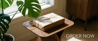 Kraft paper wrapped gift box tied with twine on wooden side table near window with monstera plant, cozy home interior, order now banner.