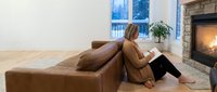 Young woman in cozy sweater reading book by fireplace in modern living room with leather sofa creating peaceful home atmosphere for relaxation and leisure.