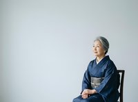 Senior Asian woman wearing traditional indigo kimono with gray obi, seated on black chair against white background, looking upward with serene expression, suitable for cultural representation, lifestyle, and wellness content.