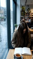 Young woman with long brown hair reading book in cozy cafe by window with tea cup on wooden table. Modern coffee shop interior with bookshelves and plants.