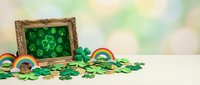 St. Patrick's Day festive display with ornate golden frame, scattered green shamrocks, rainbow decorations and gold coins on white surface against bokeh background.